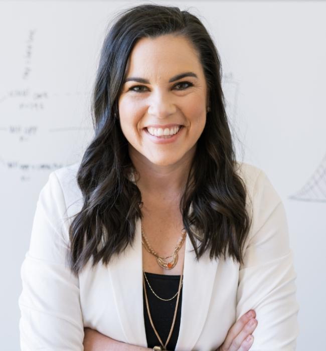 Headshot of Katelyn Jetelina, Ph.D. — a white woman with long, brown hair, standing in front of a white board wearing a white blazer and black top smiling at the camera.