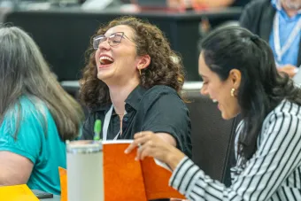 Two women sitting in a classroom at the 2025 Health Communication Leadership Institute in Austin, TX. One woman has long, curly, brown hair and is wearing glasses and a short-sleeve button down shirt. The other woman has long, black hair and is wearing a striped, long-sleeve button-down shirt.