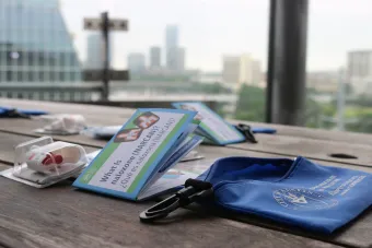 The image shows a wooden table with several items placed on it. There are pamphlets titled "What is Naloxone (NARCAN)?" in both English and Spanish, a blue pouch with the logo and text "Texas A&M University Health Science Center," and a packaged nasal spray device. The background features an out-of-focus cityscape with tall buildings.