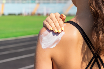 A person with yellow-painted fingernails applies sunscreen to their shoulder. The background features a blurred running track and green field, indicating an outdoor setting.