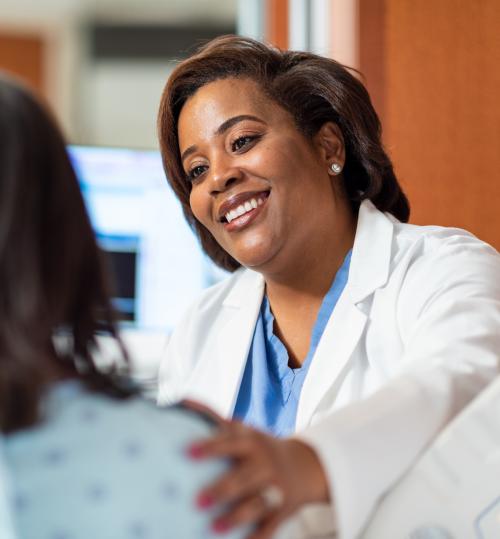 A healthcare professional in a white lab coat and blue scrubs places a hand on the shoulder of another person. Both individuals have blurred faces for privacy. Medical equipment and a computer screen displaying data are visible in the background.