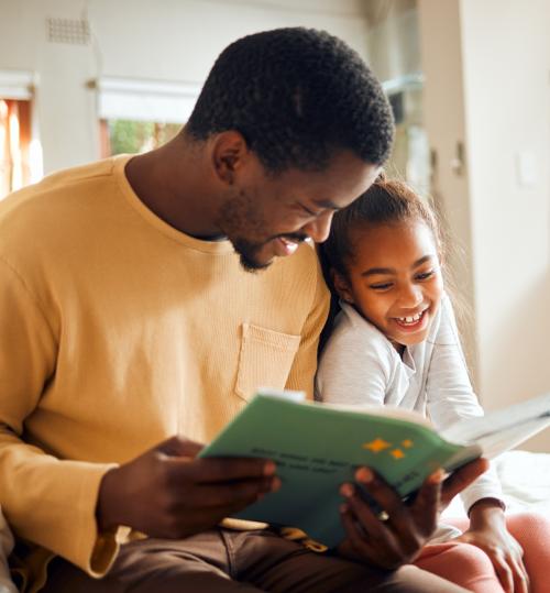 A Black man in a yellow long-sleeve shirt sits on a bed reading a book with a green cover to a young girl beside him.