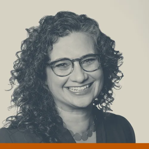 Headshot of Shana Merlin, 2025 HCLI speaker, a white woman with long curly hair wearing glasses and smiling at the camera. There is a beige and navy overlay on the photo.