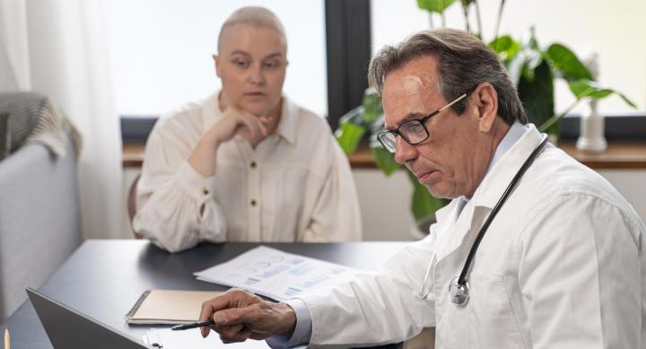 A white male doctor in a lab coat sitting at a desk and looking at a laptop with a patient.