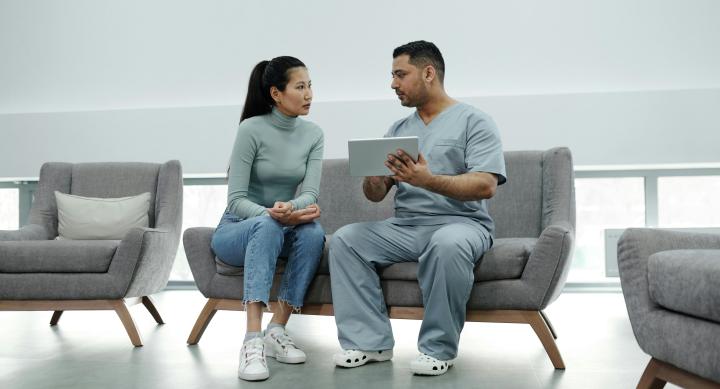 A male doctor in scrubs sitting on a couch showing a tablet to a female patient