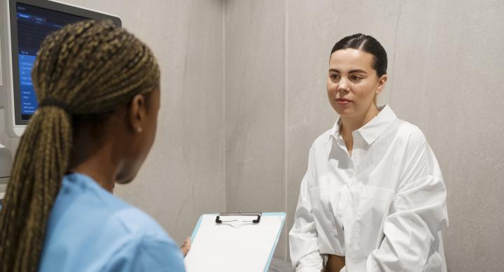 A photo of a young woman wearing a white blouse sitting in a doctor's office speaking to a nurse.