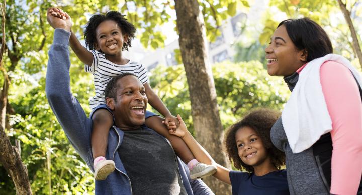 A family is enjoying time outdoors in a park. One child is sitting on an adult's shoulders, raising one arm in the air. Another child stands next to the adult, holding hands with the child on the shoulders. A second adult stands nearby, wearing a pink long-sleeve shirt and a white towel draped over their shoulder. The background features green trees and sunlight filtering through the leaves.